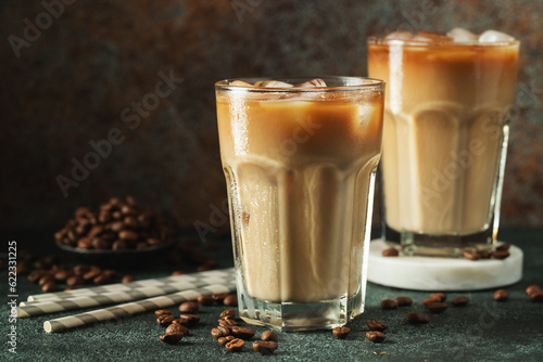 Ice coffee in a tall glass with cream poured over, ice cubes and beans on a dark concrete table.