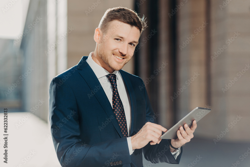 Glad confident businessman checks online bank account on touch pad, dressed elegantly, has positive expression, poses outdoors. Executive manager uses modern technologies. People and business Glad confident businessman checks online bank account on touch pad, dressed elegantly, has positive expression, poses outdoors. Executive manager uses modern technologies. People and business