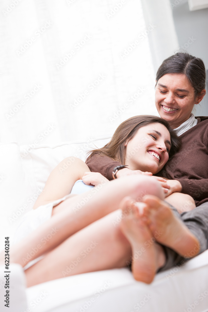 Mother and her teenage daughter relaxing together barefoot on a couch in the living room smiling with love and happiness, selective focus to their faces with copy space Mother and her teenage daughter relaxing together barefoot on a couch in the living room smiling with love and happiness, selective focus to their faces with copy space