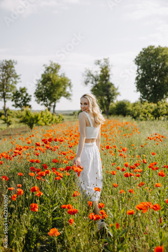 Blonde young woman standing in a field with wild red poppies, wearing a white dress, smiling, looking at camera.