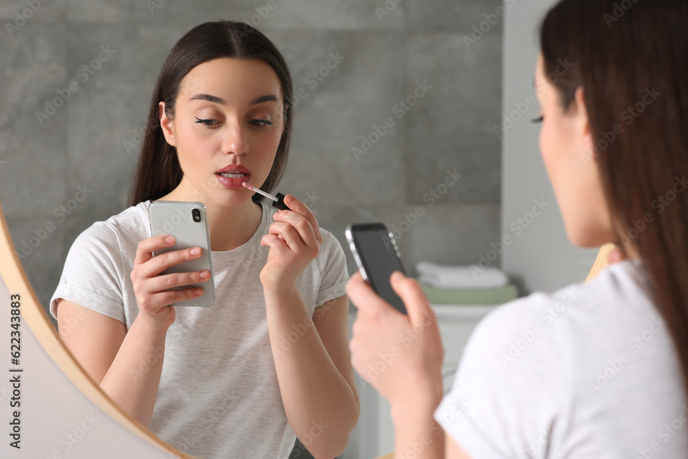Beautiful young woman using smartphone while applying lipstick near mirror indoors. Internet addiction Beautiful young woman using smartphone while applying lipstick near mirror indoors. Internet addiction