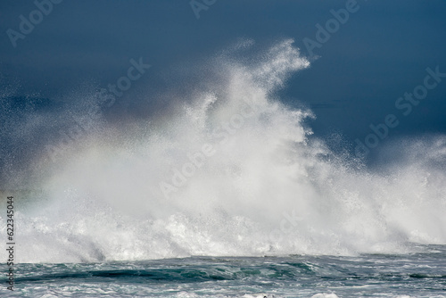 A massive wave crashes against a reef in the ocean sending water spraying high up in the air.