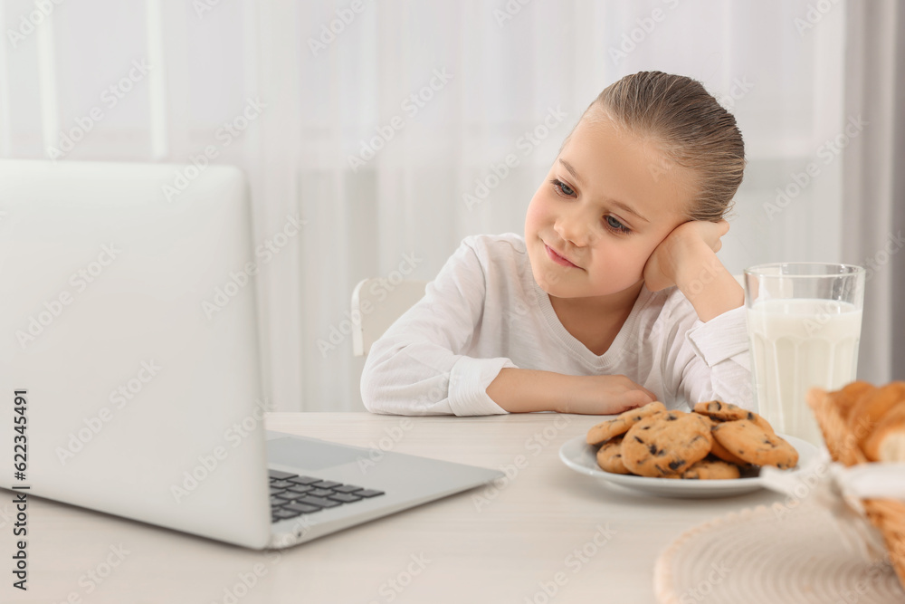 Little girl using laptop while having breakfast at table indoors. Internet addiction
