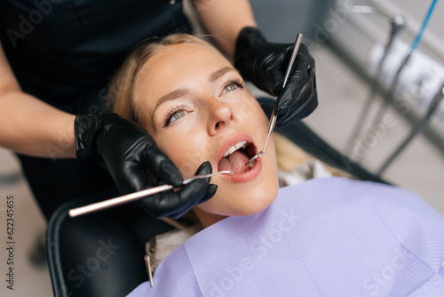 Closeup portrait of blonde female patient getting dental treatment in modern clinic, dentist doctor examining teeth of young woman with sterile tools in dentistry cabinet. Concept of dental health.