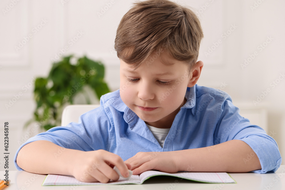 Little boy erasing mistake in his notebook at white desk indoors Little boy erasing mistake in his notebook at white desk indoors