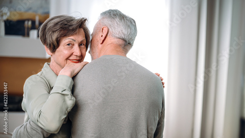 Mature smiling woman cuddling man, standing in living room