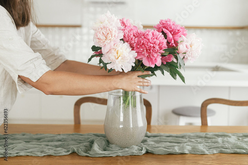 Woman arranging beautiful peonies in vase in new modern home. Young female decorating house with flowers on background of minimal white kitchen, hands with flowers close up