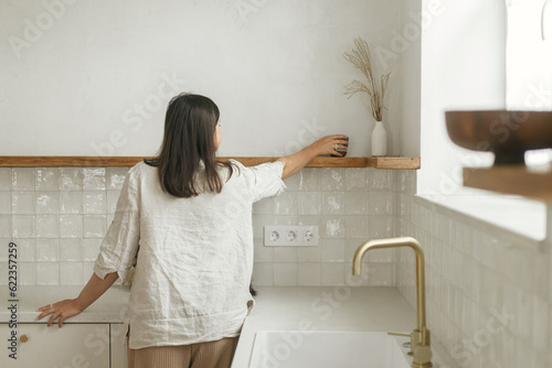Stylish woman unpacking and decorating shelves with utensils in new minimal white kitchen. Housewife cleaning up kitchen after moving in in new modern scandinavian home