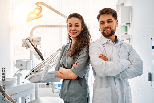 A team of doctors in a dental office standing back to back and looking at the camera and smiling. The work and leisure of doctors. In the background, a dental chair and equipment. Copy space.Sunlight. photo