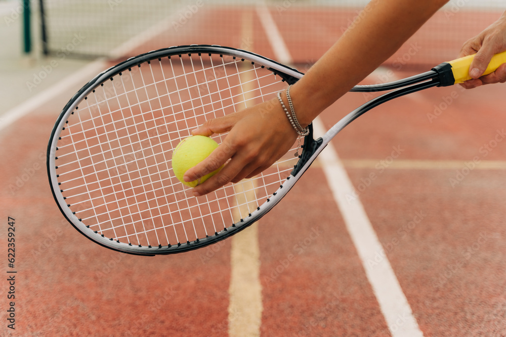 Closeup picture of attractive sporty woman hands holding yellow tennis ball and racket with tennis net and synthetic hard tennis court on the background.