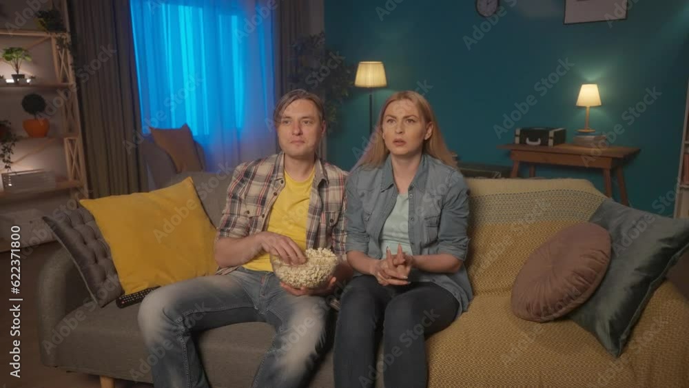 A man and a woman are watching a horror movie while sitting on the couch with a plate of popcorn. The woman is afraid, shudders, takes the man's hand and closes her eyes. HDR BT2020 HLG Material. A man and a woman are watching a horror movie while sitting on the couch with a plate of popcorn. The woman is afraid, shudders, takes the man's hand and closes her eyes. HDR BT2020 HLG Material.