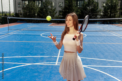 young girl tennis player in white uniform with racket throws up the ball on blue court outdoors © Богдан Маліцький