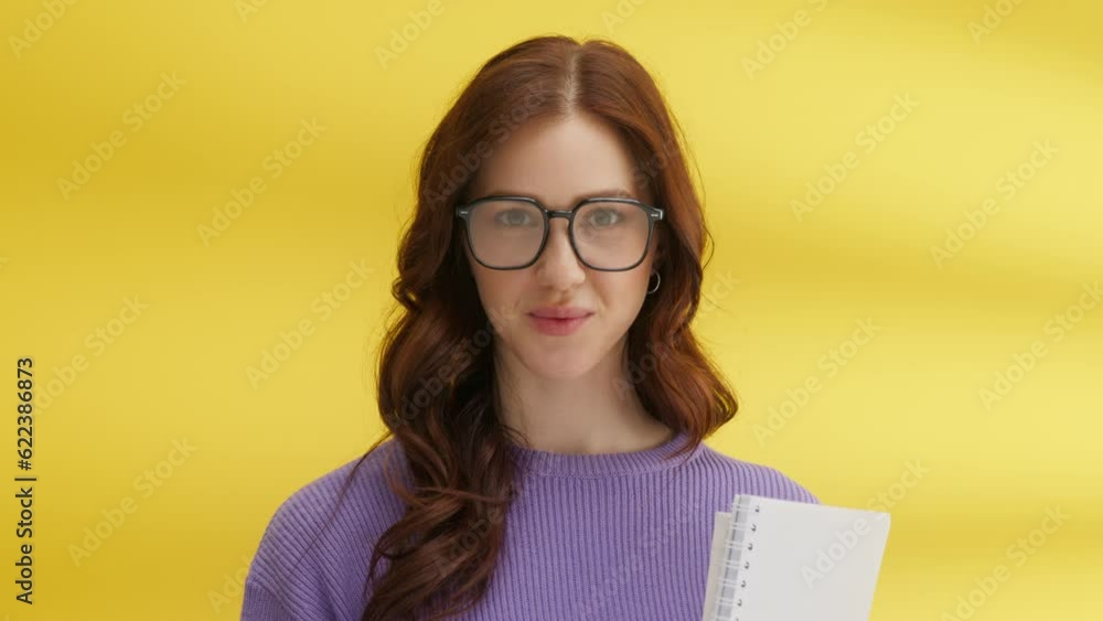 Charming long-haired brunette in big glasses, purple sweater, with slight smile, biting lip seductively, holding notebooks. Medium shot, yellow background. High quality 4k footage Charming long-haired brunette in big glasses, purple sweater, with slight smile, biting lip seductively, holding notebooks. Medium shot, yellow background. High quality 4k footage