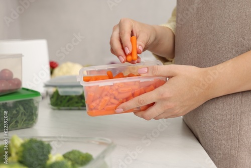 Woman putting carrots into container with fresh vegetables at white marble table in kitchen, closeup. Food storage