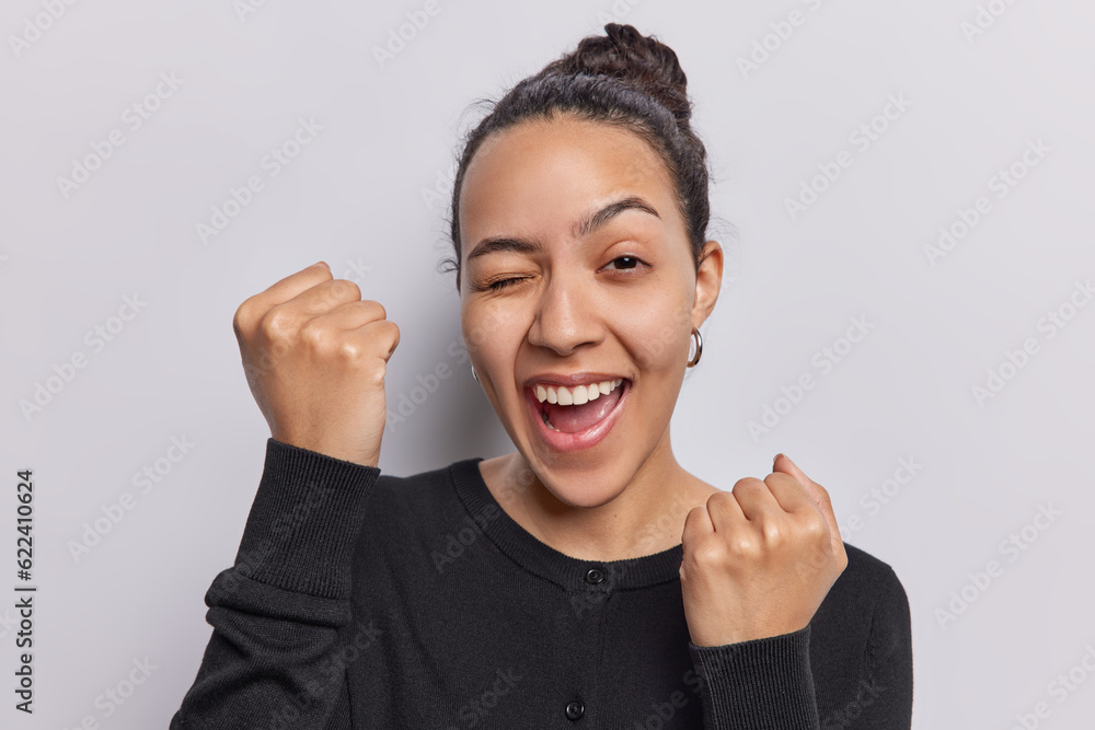 Φωτογραφία With boundless energy dark haired Latin woman wears black jumper clenches her fists exclaimes in pure joy celebrates good news while isolated on white background