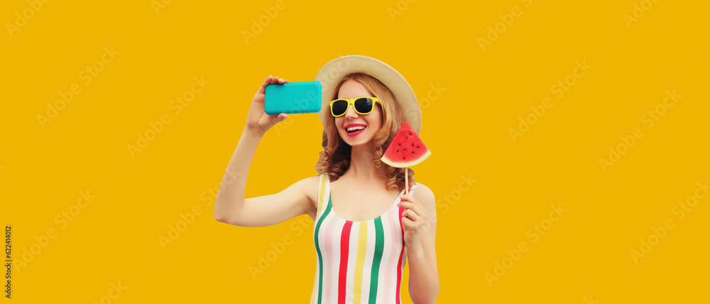 Summer portrait of cheerful happy laughing young woman taking selfie with phone and juicy lollipop or ice cream shaped slice of watermelon on orange background