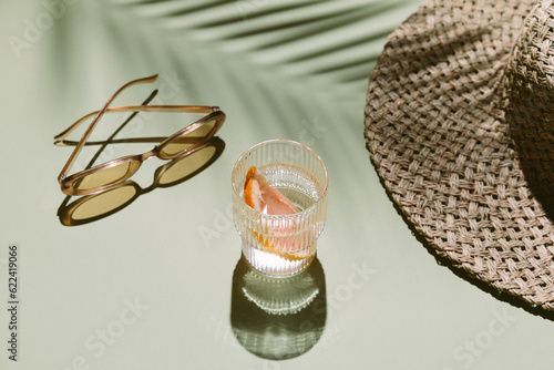 Summer flat lay with straw hat, sunglasses and glass of water with grapefruit slices on green background with palm leaf shadow, sun and sunlight. Vacation, holiday, summer creative minimal concept.