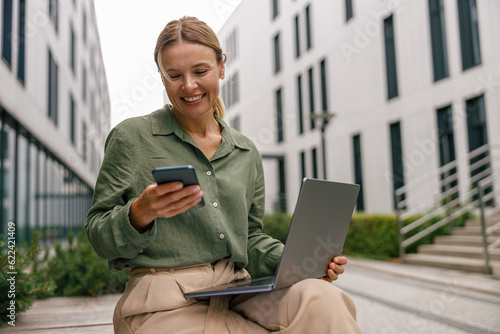 Fotografie Smiling woman manager is using phone and work on laptop outside of office