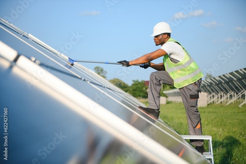 Alternative power plant worker in uniform cleaning solar panels with mop. Handsome African-American taking care of equipment.