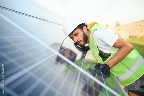 Male arabian engineer in helmet and brown overalls checking resistance in solar panels outdoors. Indian man working on station.