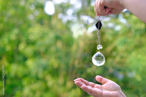 Woman hand holding and using glass ball crystal ball pendulum and using, female hands with crystal glass ball close-up on green natural background, pendulum swings, Magic and Spiritual Awakening