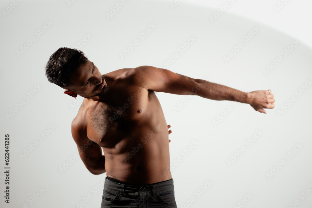 A shirtless man stretches out one arm, and tucks his other arm behind his back. He looks at his outstretched hand. Dramatic lighting adds seriousness to the bright studio setting.