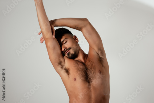 A man stretches out his arms and leans to the side, with his eyes closed. He is shirtless, and he looks confident and comfortable in his own skin and in his body.