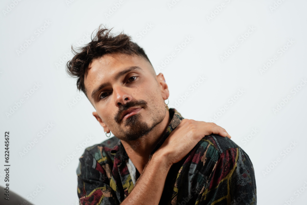 A handsome man rests his hand on his shoulder and leans to the side. He has a goatee and wears earrings, alluding to his vibrant style. A handsome man rests his hand on his shoulder and leans to the side. He has a goatee and wears earrings, alluding to his vibrant style.