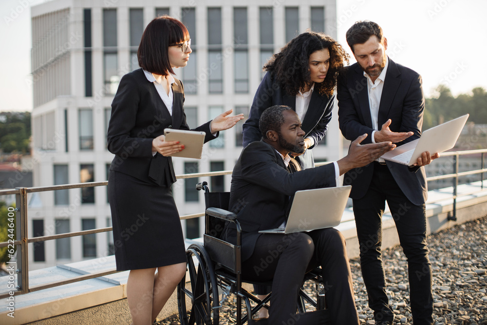 Multiracial team of company representatives with man on wheelchair making business appointment on office rooftop. Experienced colleagues holding digital gadgets and disputing about project financing. Multiracial team of company representatives with man on wheelchair making business appointment on office rooftop. Experienced colleagues holding digital gadgets and disputing about project financing.