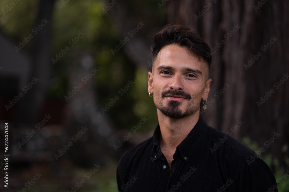 With a gentle smile, a handsome man turns to look at the camera. He has a fresh haircut, trimmed goatee, and delicate earrings. He is standing in front of an enormous, old redwood tree in a park. With a gentle smile, a handsome man turns to look at the camera. He has a fresh haircut, trimmed goatee, and delicate earrings. He is standing in front of an enormous, old redwood tree in a park.