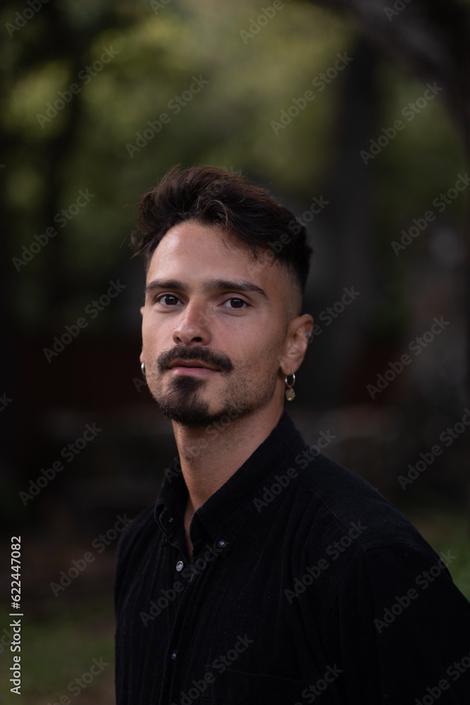 A handsome man with a goatee, earrings, and a fresh haircut looks over to pose for a serious portrait. He is wearing a black shirt, and a redwood forest is behind him.