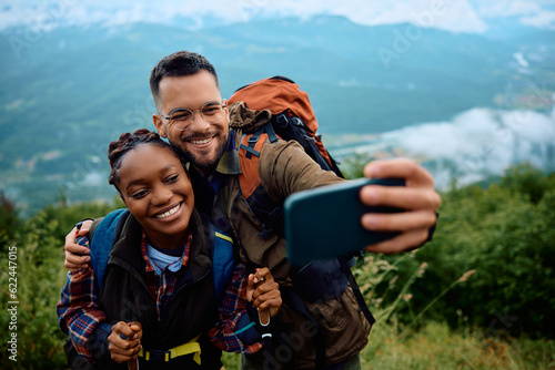 Happy hiker and his black girlfriend taking selfie with cell phone on top of hill.