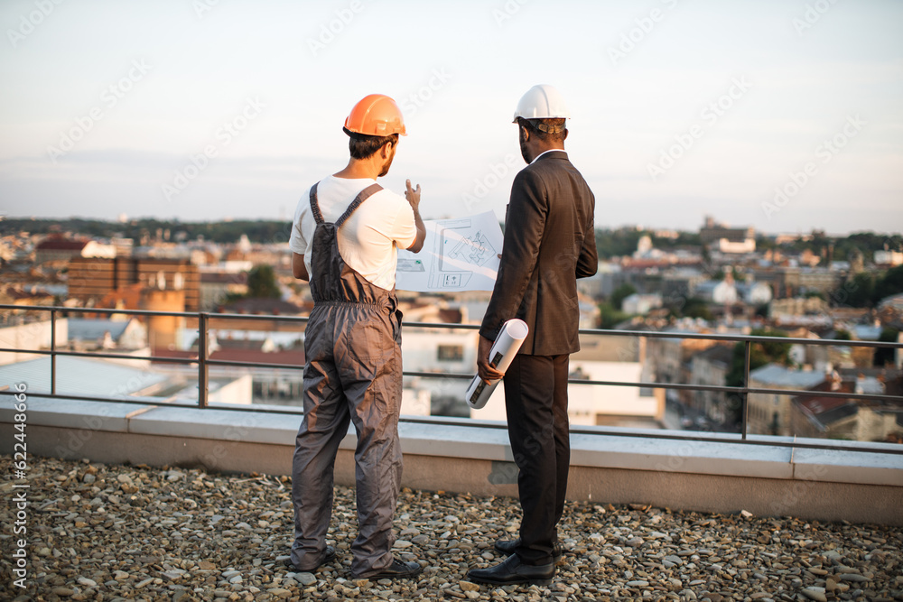 Back view of two male builders in protective helmets watching at construction site from rooftop of city building. Foreman explaining details from technical drawings to african american engineer.