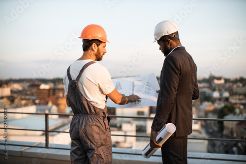 Rare view of caucasian bearded workman presenting design blueprints to african american urban planner on fresh air. Male developers in hardhats discussing location of balconies on apartment building.