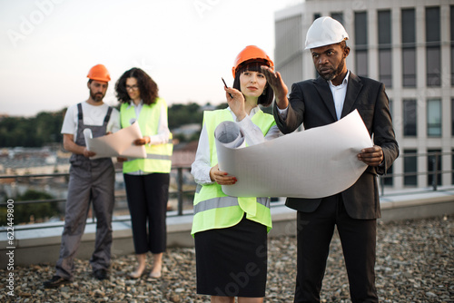 Group of diverse engineers with blueprints standing on flat rooftop and talking about construction process. Two colleagues of caucasian and african american nationality pointing at building site.