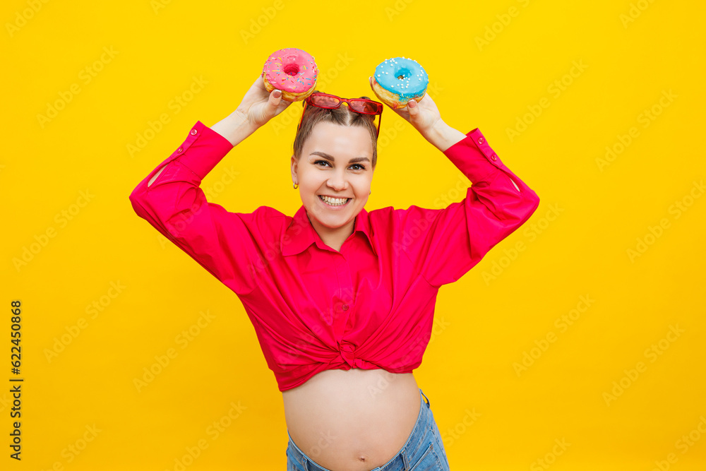A beautiful pregnant woman holds a sweet donut in her hand on a yellow background. Expecting a child, pregnancy and motherhood. The concept of healthy and unhealthy food, diet. Junk food A beautiful pregnant woman holds a sweet donut in her hand on a yellow background. Expecting a child, pregnancy and motherhood. The concept of healthy and unhealthy food, diet. Junk food