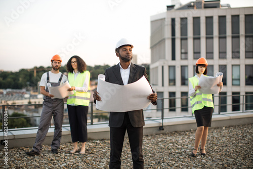 African american businessman wearing suit and hardhat holding project plan in hands and looking aside. Multicultural developers watching at blueprints and discussing construction details.