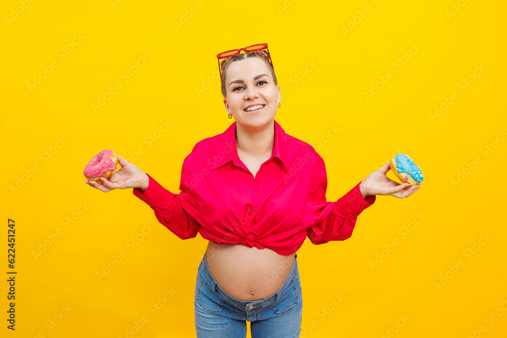 A smiling pregnant woman in a pink shirt holds donuts in her hands on a yellow background. Sweet food during pregnancy. Harmful food during pregnancy. A smiling pregnant woman in a pink shirt holds donuts in her hands on a yellow background. Sweet food during pregnancy. Harmful food during pregnancy.