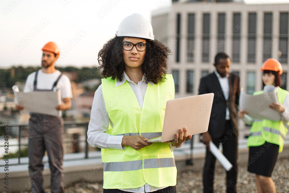 Portrait of african american female urban planner in protective wear and glasses holding remote computer and looking at camera. Diverse architects reviewing building blueprints on roof terrace.