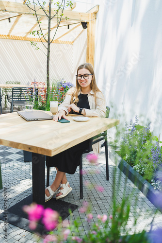 Young European woman sitting at outdoor cafe table with laptop and cup of coffee, smiling woman in glasses enjoying telecommuting in cafe or studying online
