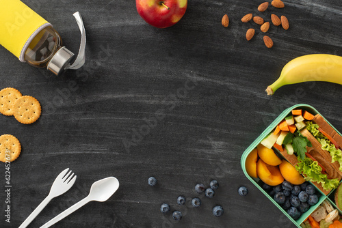 Creative school snack concept. Top view of plastic lunchbox showcasing colorful assortment of nutritious foods, water bottle, cutlery. Set against blackboard backdrop, perfect for text or promotions