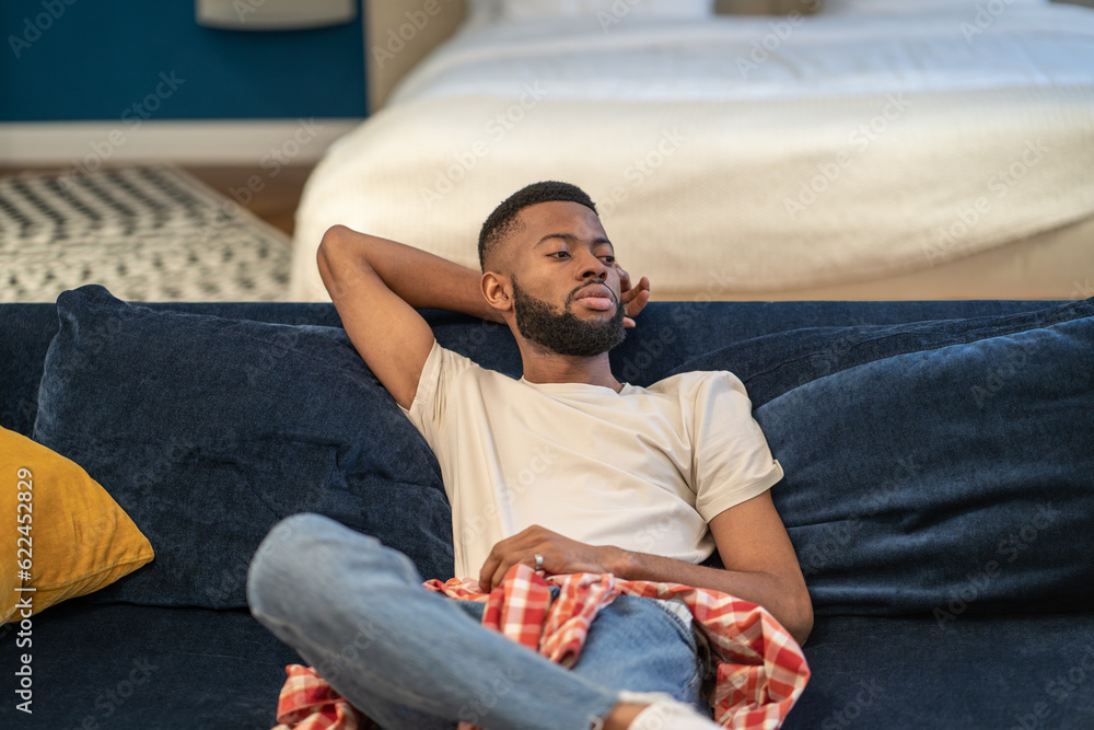 Fototapeta premium Tired pensive black man relaxing on comfortable couch enjoying lazy weekend at modern interior home. . Thoughtful bearded dreaming African American guy sits on sofa rests in studio apartment.