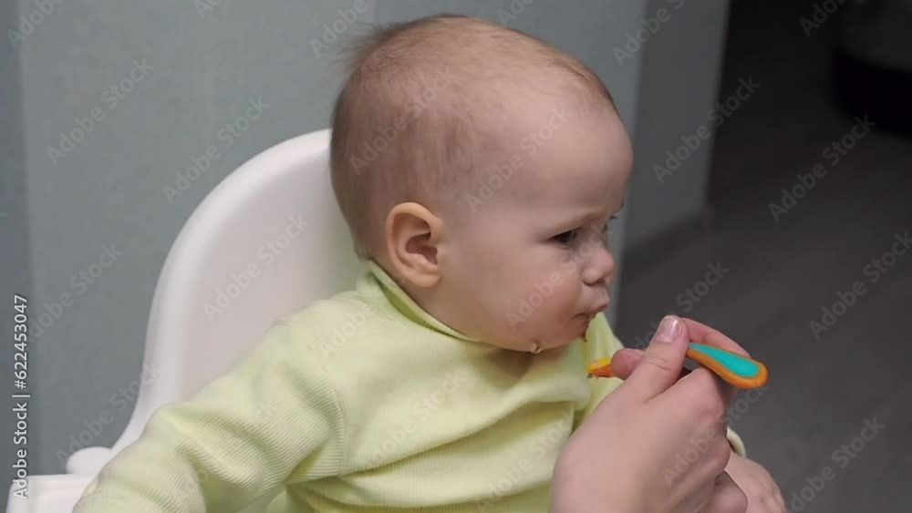 Mother feeding her daughter baby girl with spoon in baby chair. Child with appetite to eat porridge on kitchen Mother feeding her daughter baby girl with spoon in baby chair. Child with appetite to eat porridge on kitchen