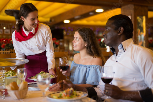 Smiling waitress serving food for positive couple at restaurant, putting plate with salad on table