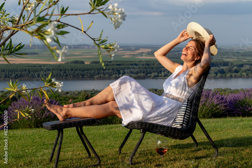 A woman in a white dress and a straw hat with a glass of wine enjoying life on a sunny afternoon outdoors.