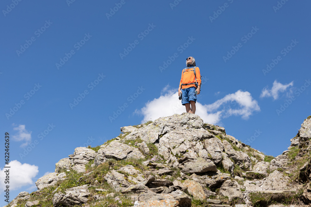 A man with a backpack stands on a rocky hill against a bright blue sky, Austria