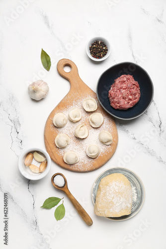 Wooden board with uncooked dumplings and ingredients on white marble background