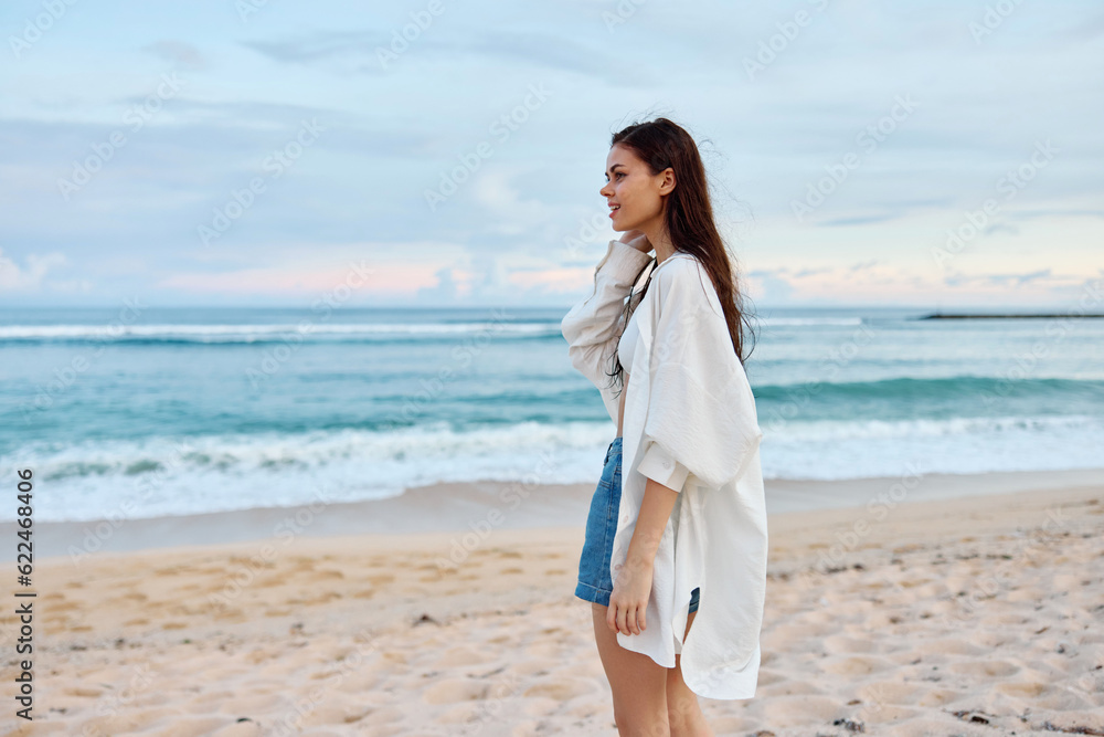 Happy tanned slim woman in white swimsuit shirt and jean shorts walks on the beach on the sand by the ocean with wet hair after swimming, sunset light