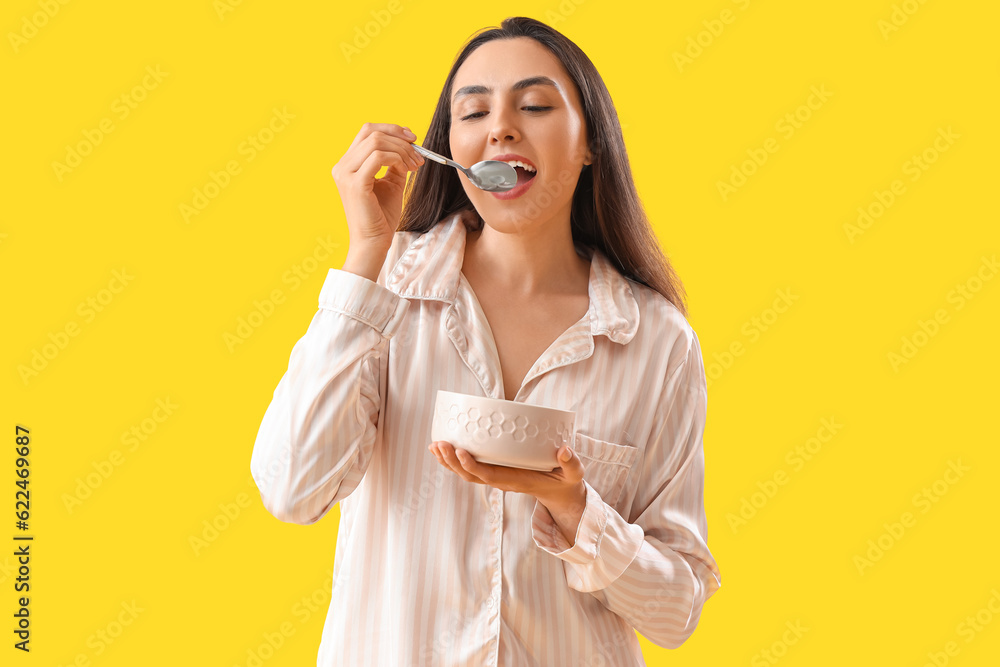 Young woman in pajamas with spoon and bowl on yellow background Young woman in pajamas with spoon and bowl on yellow background