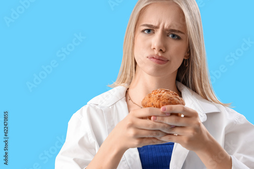 Worried young woman with tasty muffin on blue background, closeup